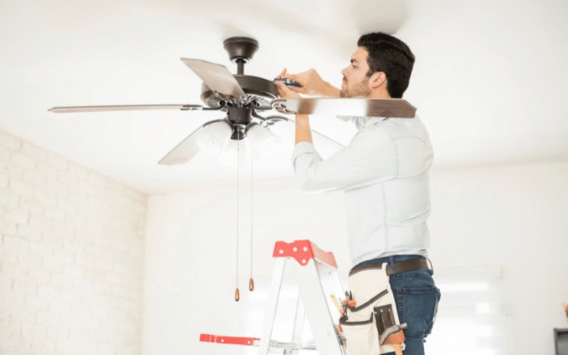 A handyman Davidson & Williamson County stands on a ladder using a screwdriver to install or repair a ceiling fan, with various tools in his belt, in a bright, modern TN room.