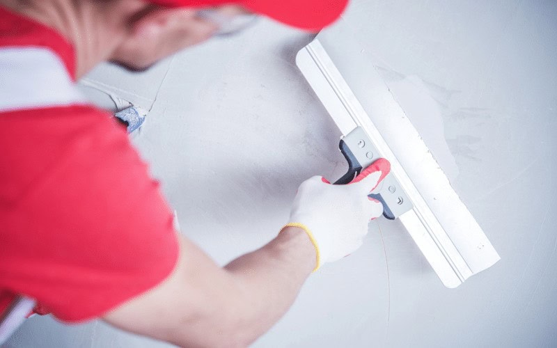 A handyman in Davidson & Williamson County, TN, wearing a red shirt and white gloves, smooths a wall with a large drywall taping knife, carefully applying or spreading plaster.