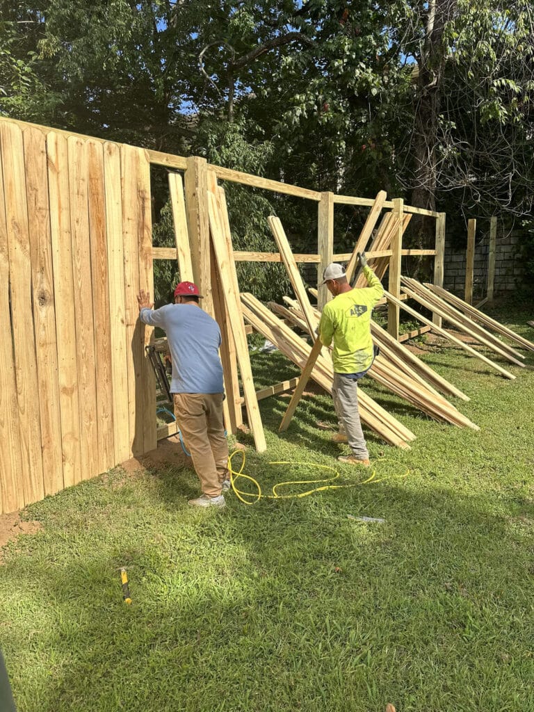 Two workers from a handyman Davidson & Williamson County, TN team install a wooden fence in a grassy yard. One holds a plank steady while the other manages more planks, with stacks of lumber nearby and trees in the background.