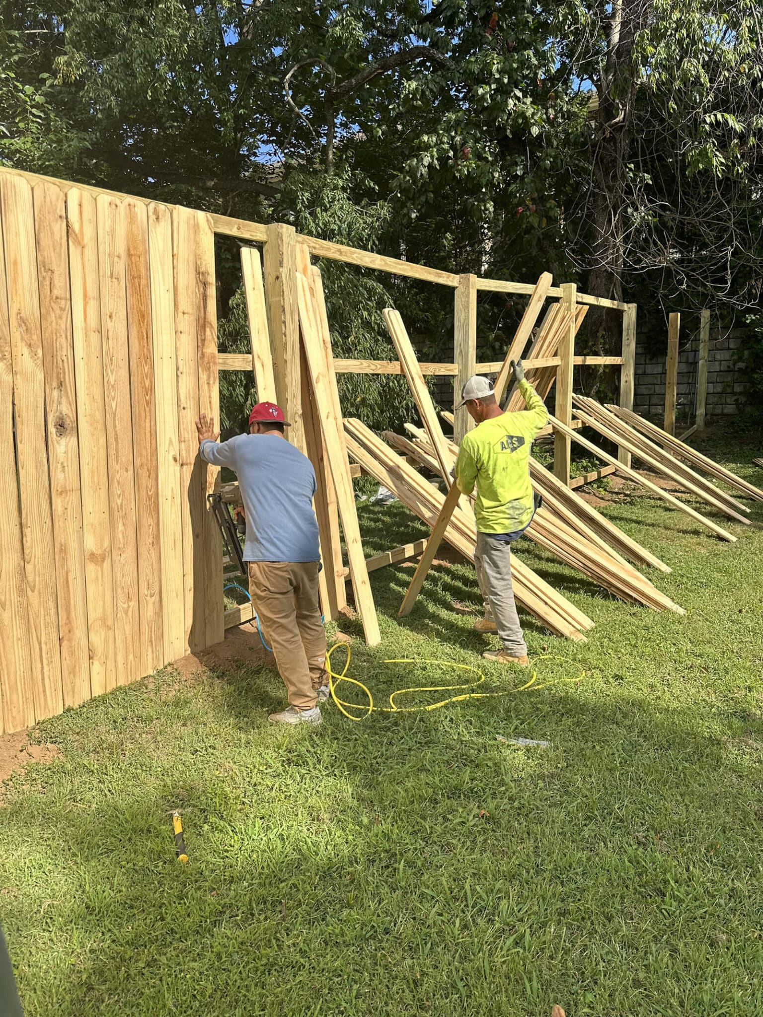 Two workers from a handyman Davidson & Williamson County, TN team install a wooden fence in a grassy yard. One holds a plank steady while the other manages more planks, with stacks of lumber nearby and trees in the background.