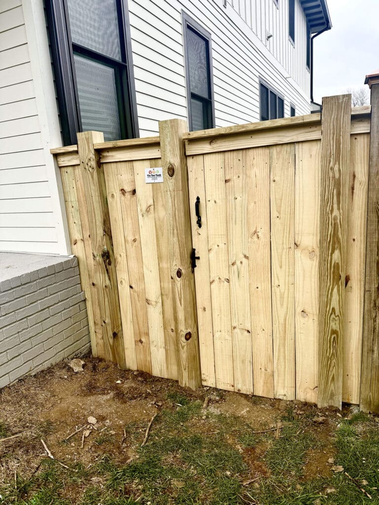 A wooden fence with a gate stands next to a white house in TN. The fence has vertical planks, a black metal latch, and a small sign near the top—expertly crafted by a handyman Davidson & Williamson County residents trust. Grass and bare soil are visible in the foreground.