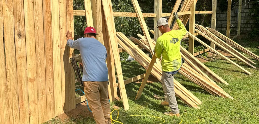Two construction workers are installing wooden boards to build a privacy fence in a grassy yard. If you need a skilled handyman in Davidson & Williamson County, TN, they can help with fencing projects and more. Stacks of boards lean against the fence frame.