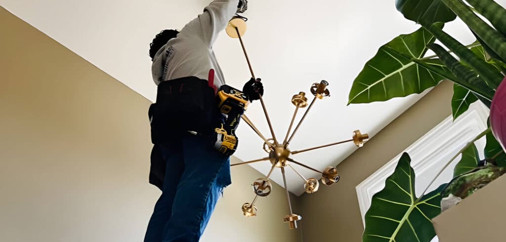 A handyman in Davidson & Williamson County, TN stands on a ladder, installing a modern gold chandelier with a power drill. Large green houseplants and natural light from a window brighten the scene.
