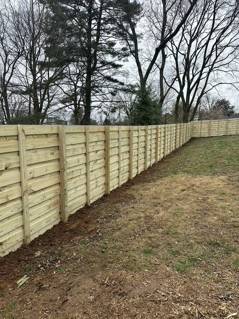 A newly built wooden privacy fence stretches across a grassy TN yard, with tall trees and bare branches visible in the background under a cloudy sky—another quality project by a handyman Davidson & Williamson County trusts.