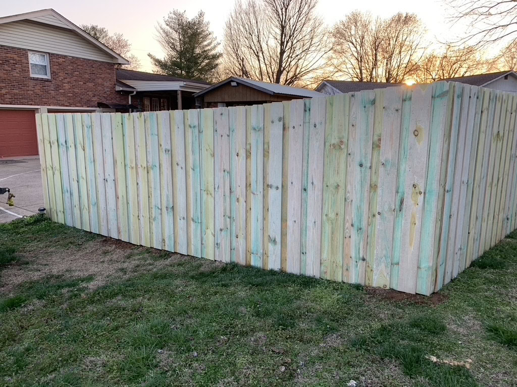 A newly installed wooden fence with greenish stains stands in a suburban yard. Houses and trees are visible in the background at sunset, expertly crafted by a handyman in Davidson & Williamson County, TN.