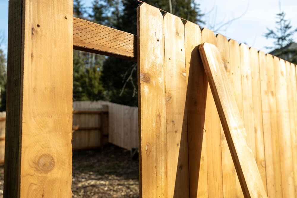 Close-up of a wooden fence under construction by a handyman in Davidson & Williamson County, TN, with one section missing a panel and a diagonal support beam leaning against the fence. Trees and more fencing are visible in the background.