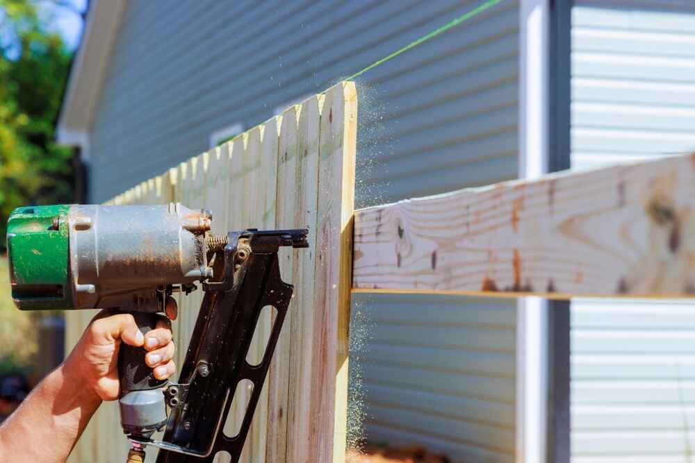 A handyman in Davidson & Williamson County, TN uses a nail gun to attach wooden fence boards to horizontal supports, with a house and siding visible in the background.