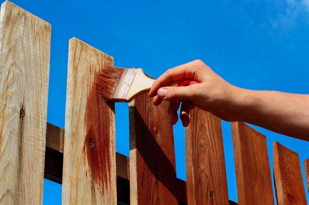 A handyman in Davidson & Williamson County, TN is painting a wooden fence with a brush, applying brown wood stain to the vertical boards under a clear blue sky.
