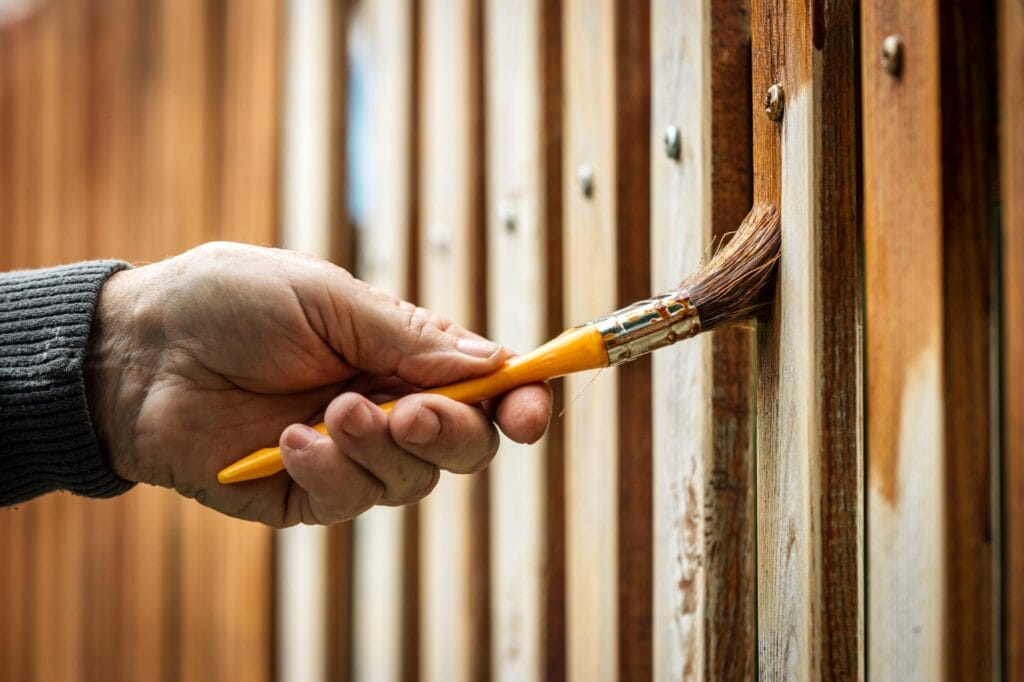 A person’s hand, possibly a handyman Davidson & Williamson County, holds a yellow paintbrush painting weathered wooden fence planks. The wood looks aged, and the person is wearing a gray long-sleeve sweater in TN.