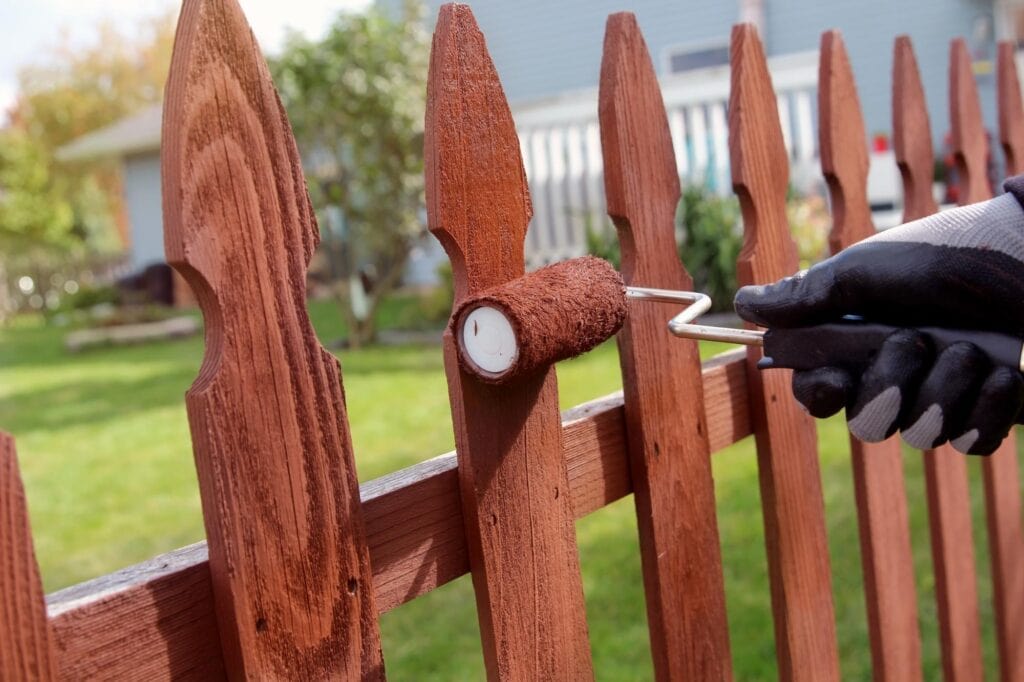 A gloved hand uses a paint roller to apply brown stain to a wooden picket fence in a sunny backyard. Green grass and a house are visible in the background—perfect handiwork from a handyman in Davidson & Williamson County, TN.