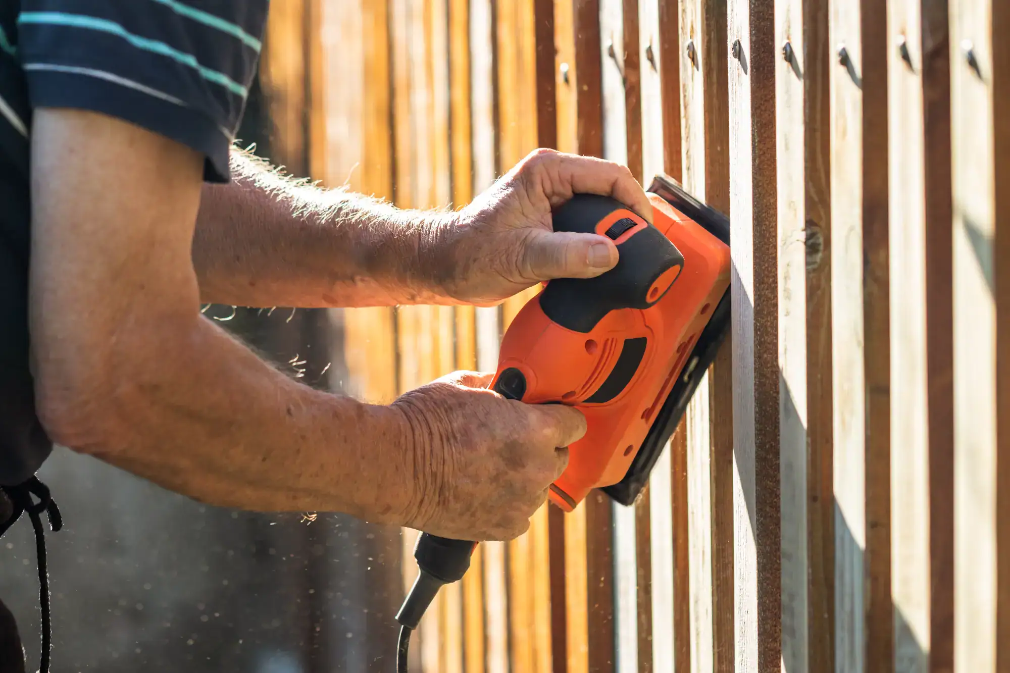 Close-up of a person using an orange electric sander to smooth a wooden fence outdoors in TN, with sunlight illuminating the wood and dust particles, showcasing the handiwork of a handyman Davidson & Williamson County expert.