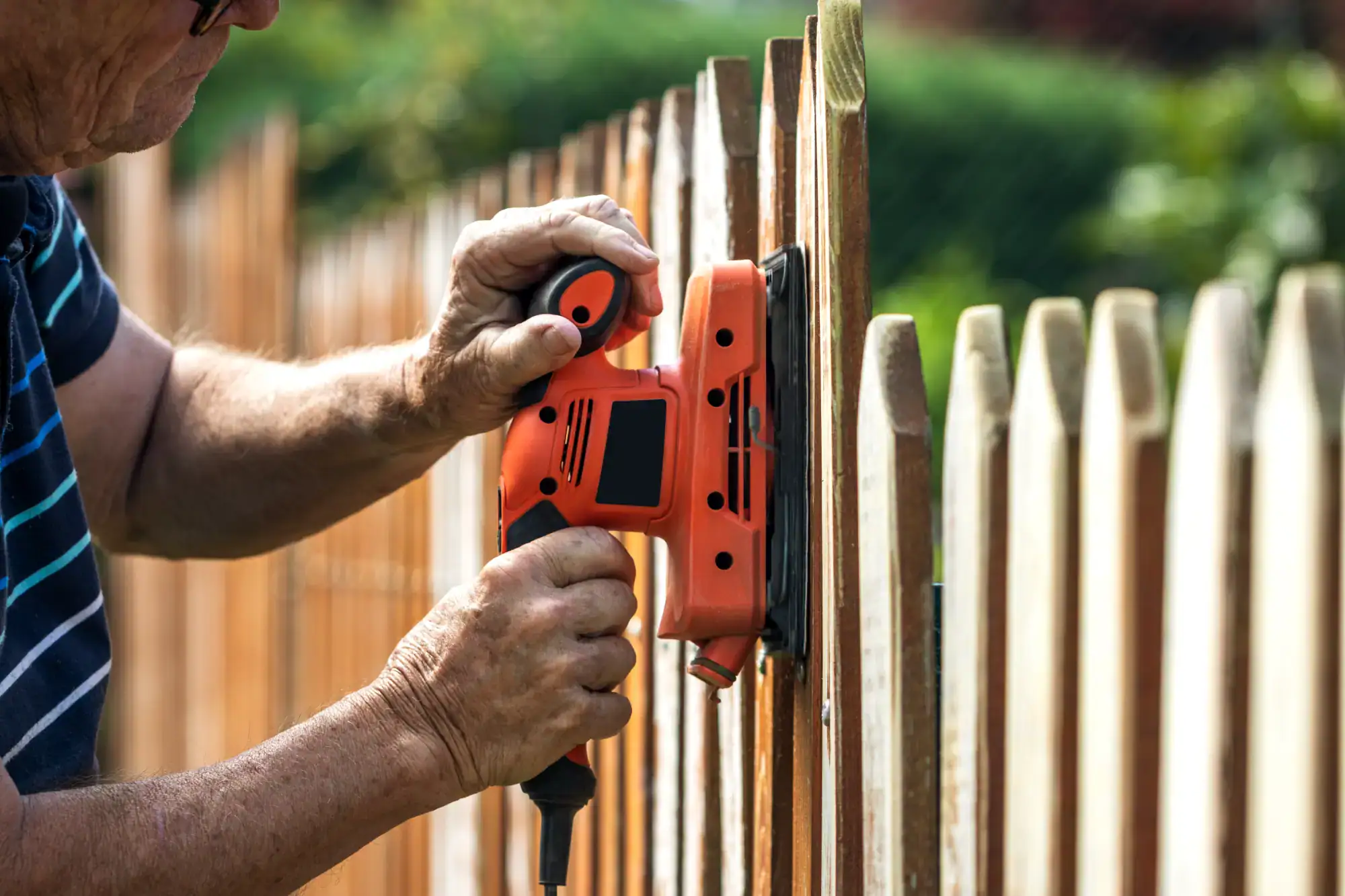 A handyman in Davidson & Williamson County, TN uses an orange electric sander to smooth the wooden pickets of a fence outdoors on a sunny day.