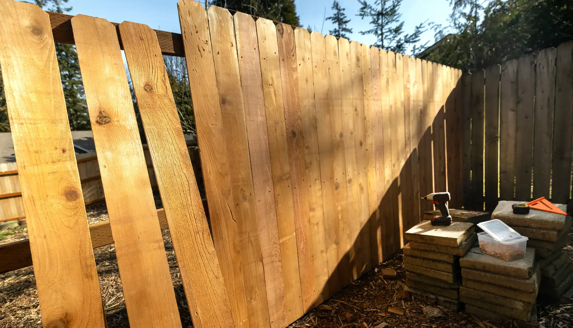 A wooden fence is partially constructed in a yard by a handyman in Davidson & Williamson County, TN. Tools, a drill, and supplies rest on stacked concrete pavers nearby as sunlight casts shadows on the fence amid trees in the background.
