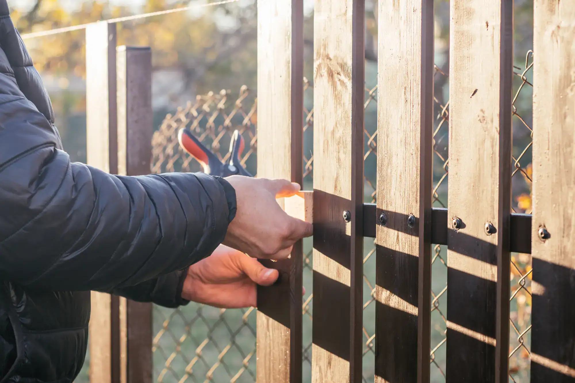 A handyman in Davidson & Williamson County, TN, wearing a dark jacket, uses a screwdriver to secure wooden planks to a fence outdoors, with sunlight and a wire fence visible in the background.